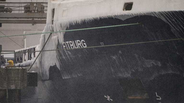 A ship with "Fitburg" written on its side, docked at the harbor. There are large icicles on the side of the ship.