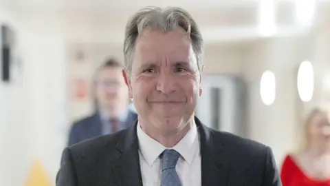 PA Media A photograph of the MP Dan Norris wearing a suit and tie. Two people who are not in focus can be seen in the background 