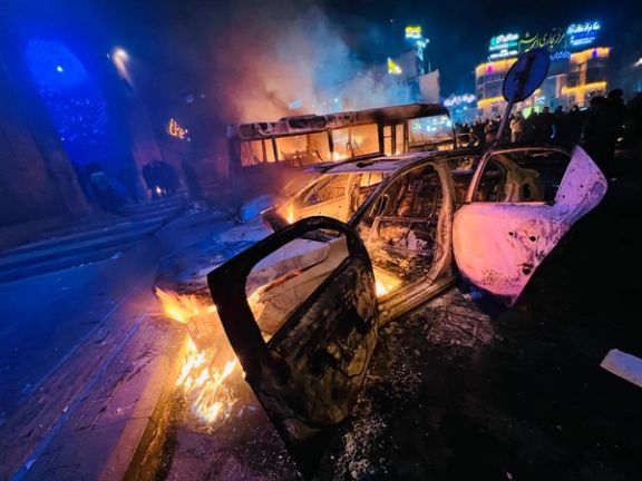 A burned-out car and bus continue to smolder in Saadatabad in northern Tehran on January 10, as crowds gather nearby during an overnight protest.