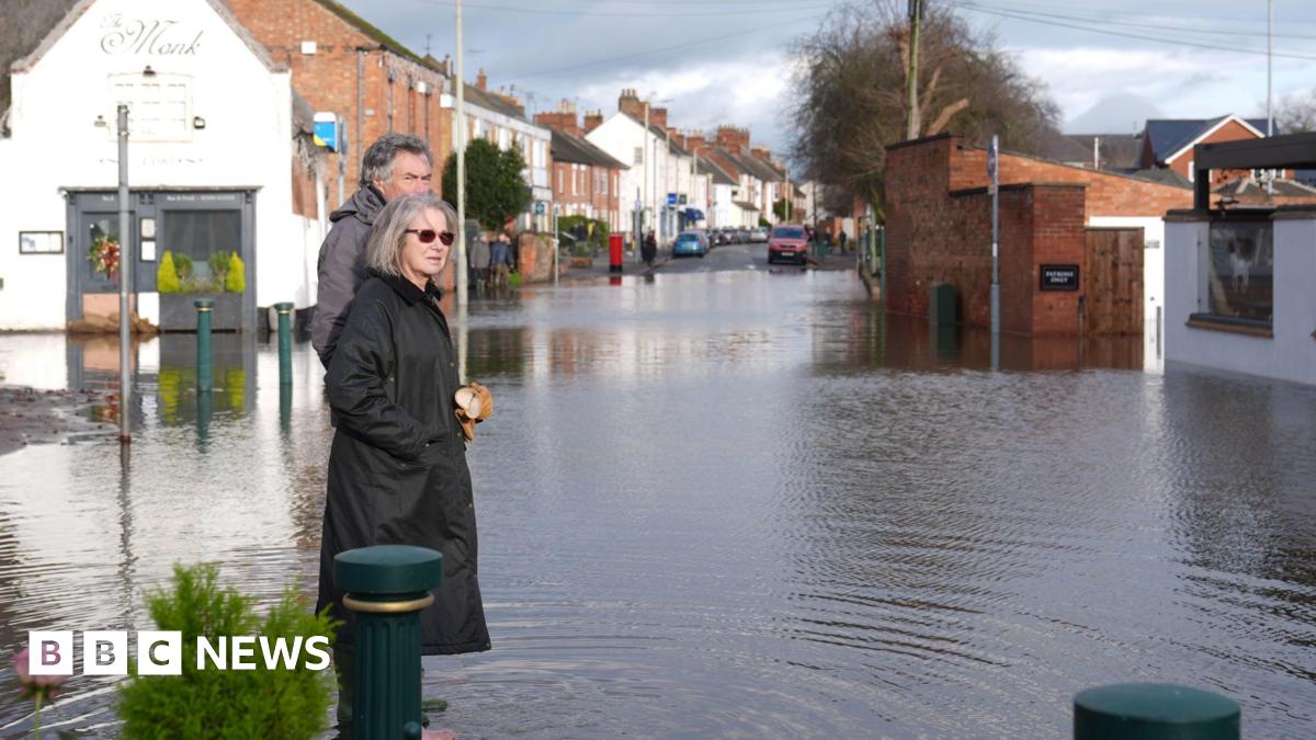 A man and a woman, both in winter coats, survey the flood waters in Station Road, Quorn, which is around ankle depth. Ripples can be seen in the water and the Monk restaurant partly submerged in the background.