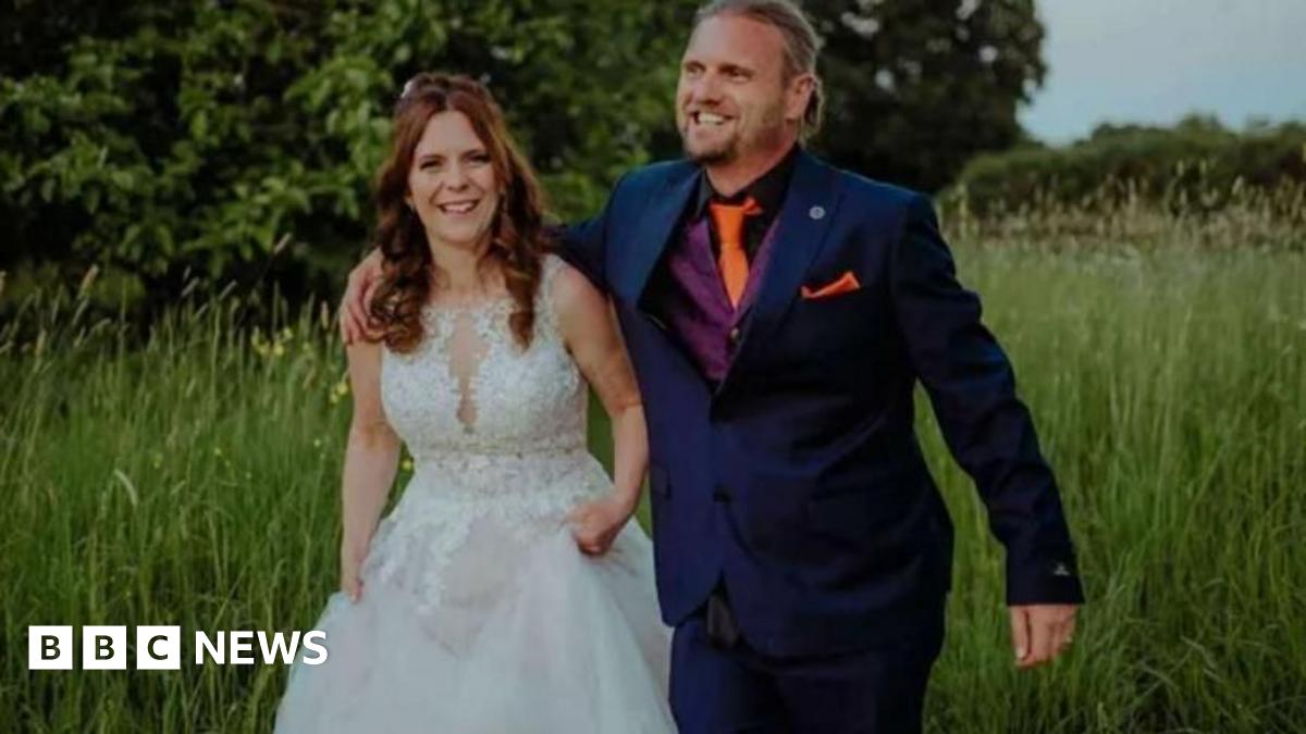 Kayleigh and Christopher Coll walking through a field of long grass and smiling - Mrs Coll is wearing a white wedding dress and Mr Coll is wearing a dark blue suit, purple waistcoat and orange tie.