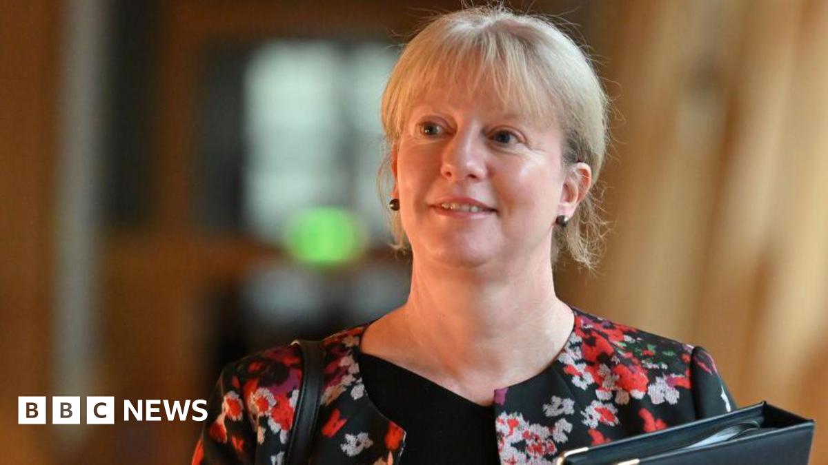 Shona Robison, with blonde hair and wearing a black top and patterned jacket, walks in the Scottish Parliament, carrying a black folder.