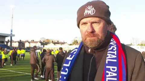 Anthony Dearnley is wearing a beige-coloured woollen hat and a commemorative match scarf and brown top and jacket is standing next to the pitch.