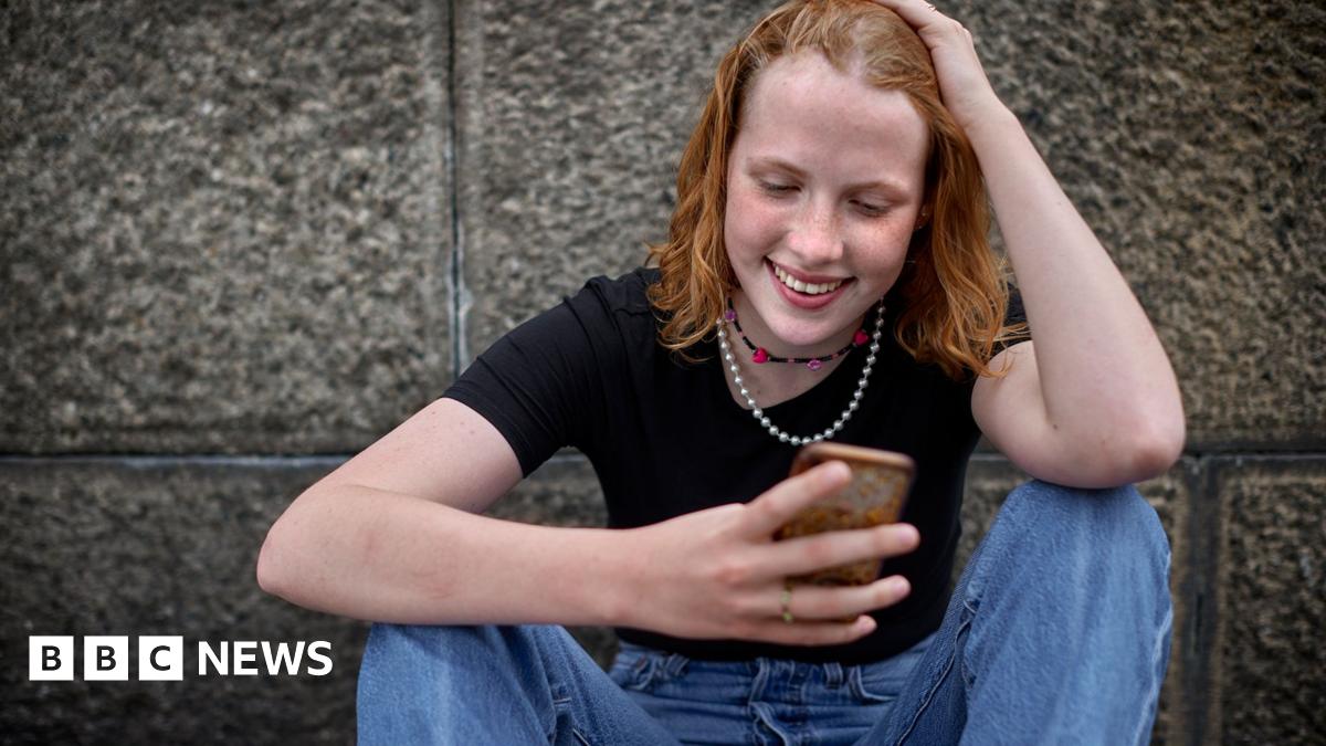 A girl looks at her phone, she has her hand on her head smiling and is wearning a black top and blue jeans.