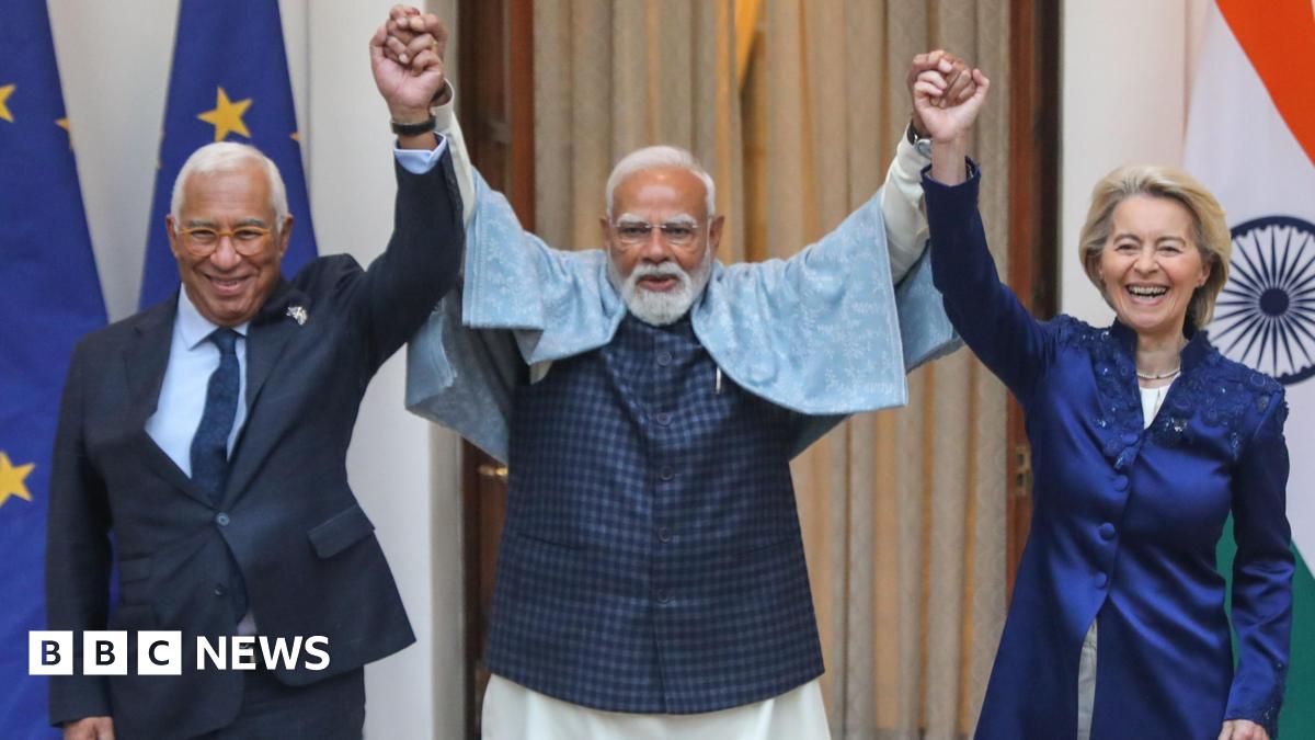 Three leaders hold hands and raise their arms, laughing during a friendly moment. The man in the centre, Indian Prime Minister Narendra Modi, with a white beard and glasses, wears a light shawl over a dark outfit. On either side, European Council President António Costa in a dark suit and European Commission President Ursula von der Leyen in a dark blue long jacket