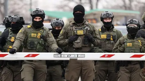 Getty Images US Border Patrol agents stand behind a barrier. Their faces are covered and they are all wearing tactical vests and olive green overalls.