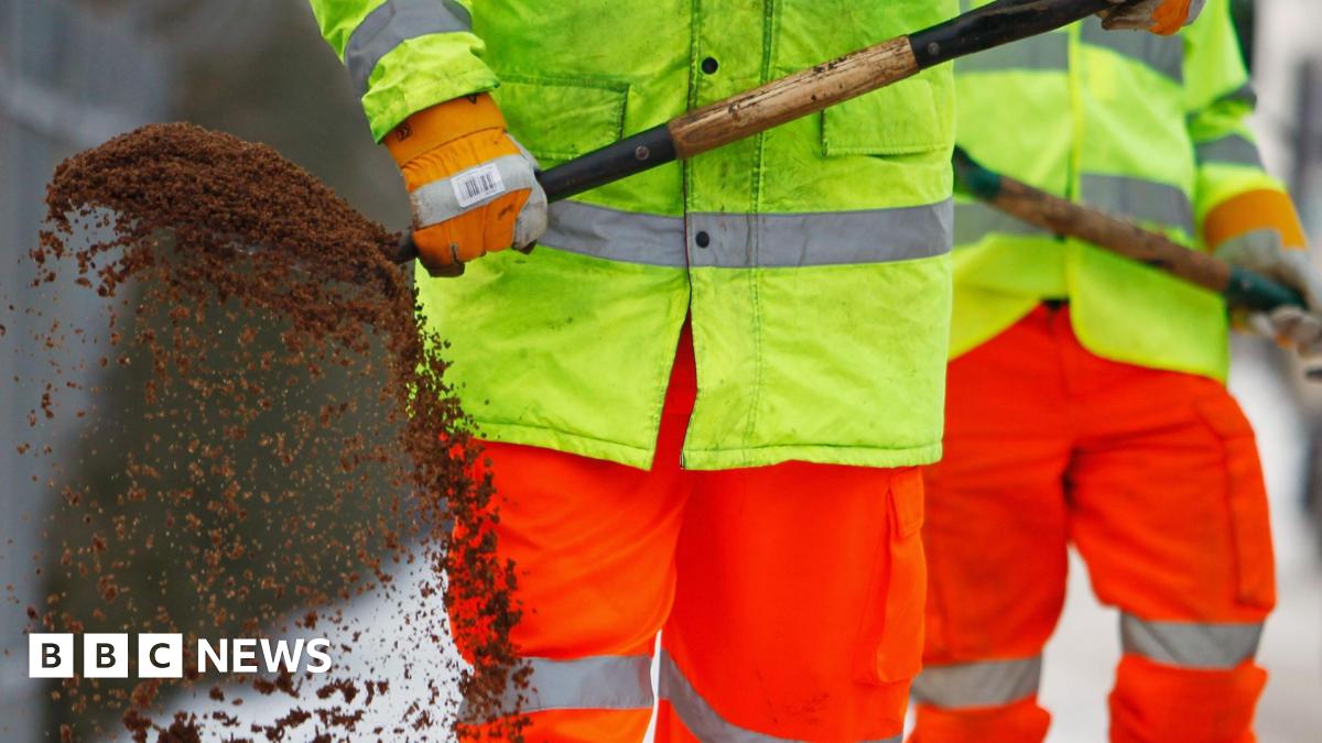 Millie Bobby Brown and David Harbour in character as Eleven and Jim Hopper. Eleven wears red shorts over grey leggings and a grey jumper. Jim Hopper wears a black leather jacket, a black beanie hat and dark trousers.They are speaking to director Frank Darabont on the set of Stranger Things. There is a vintage advertisement posted in the background with debrief on the ground.