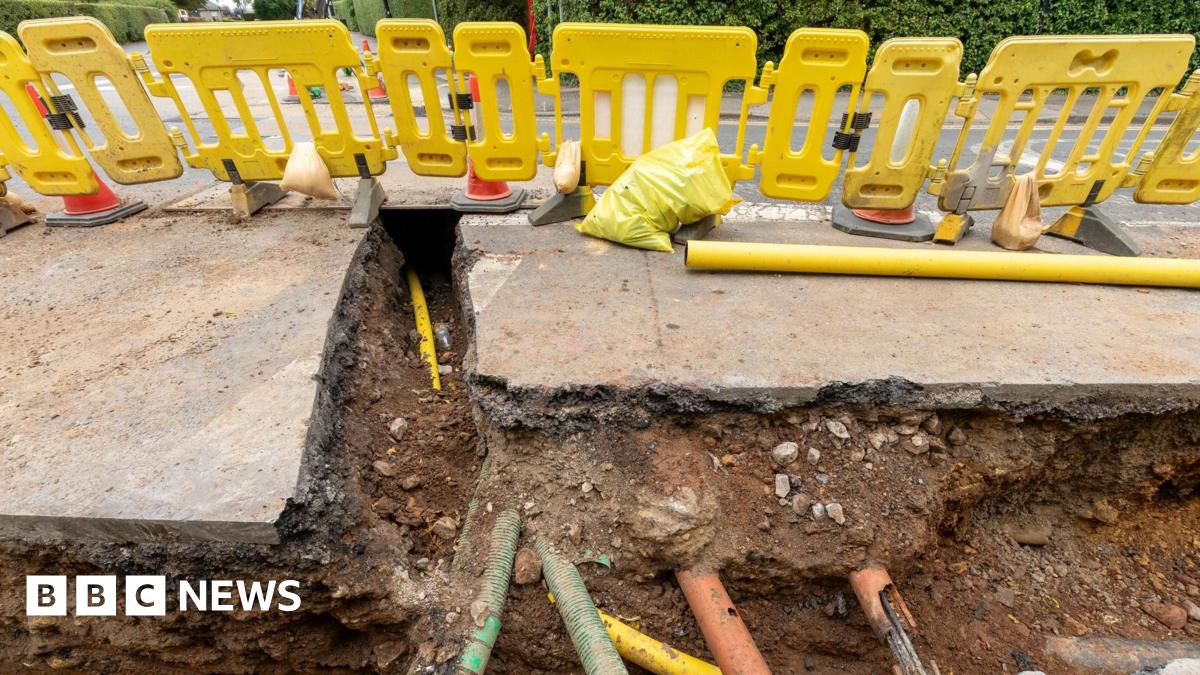 A hole in a road  surrounded by yellow safety fencing, with orange, yellow and green pipes visible underground.