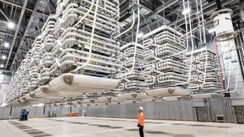 National Grid A man looks up at machines made up of thick white pipes with silver boxes inside hanging from the ceiling inside the valve hall. The hall is situated within the converter station, which is part of the onshore infrastructure associated with an interconnector. And it is where the conversion between DC and AC takes place.