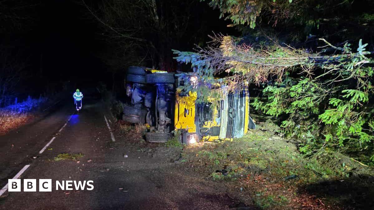 A gritter lorry on its side on a roadside in the early hours. It is dark and there is lighting on it.