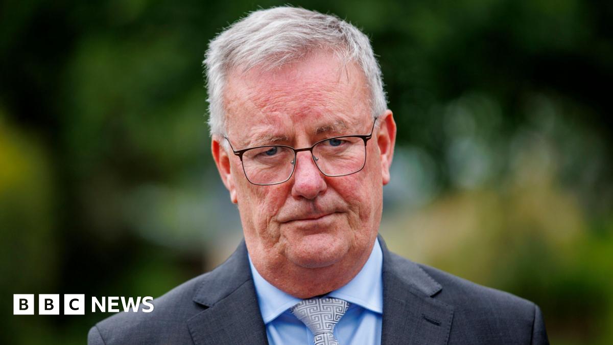 A man is looking into the camera. He has a neutral expression. He's wearing glasses and a grey suit with a blue shirt and a grey tie with a maze design. He's outside. Trees in the background are out of focus.