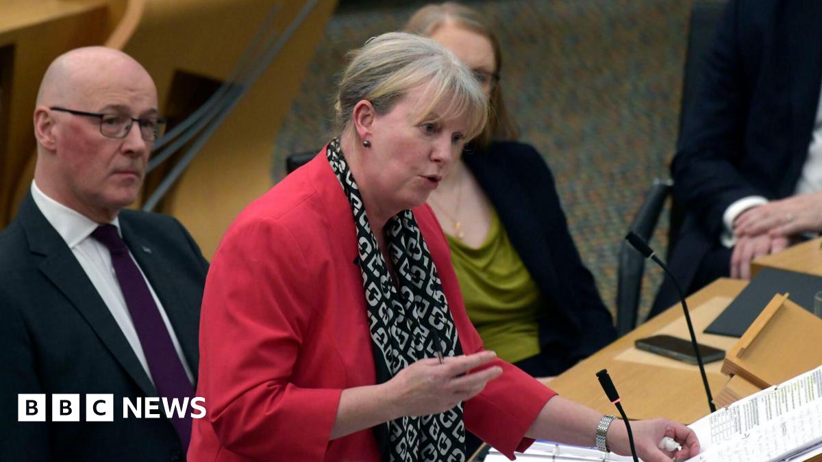 Finance Secretary Shona Robison announces the draft Budget for 2026-27 to MSPs at the Scottish Parliament in Holyrood. She stands in the debating chamber at a desk with two folders in front of her. She has short, tied back fair hair and wears a pink top and black and white scarf and is speaking. John Swinney sits next to her and looks on, as do other MSPs in the background.