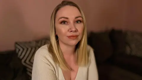 Martin Giles/BBC Abbie looking into camera. She is sat on a sofa with cushions behind her. She has blonde hair and is wearing a beige jumper.