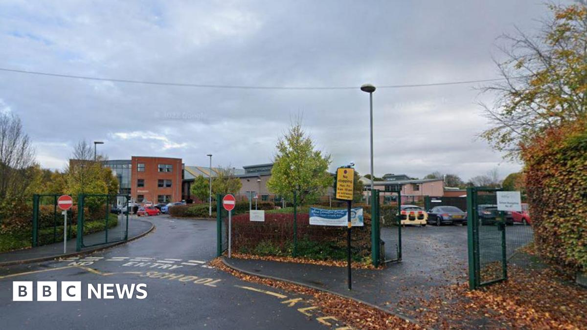 the outside of a secondary school, pictured from gates by its entrance. Green metal fencing bars an entrance and exit and a carpark can be seen in front of buildings. Hedges and trees line the perimeter. The school buildings can be seen in the distance, with three storeys. The sky is grey and cloudy.