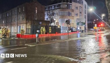 Water flows down a road.  A handful of firefighters stand behind an orange barricade