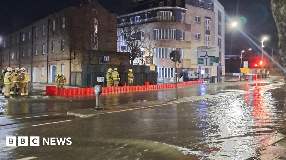Water flows down a road.  A handful of firefighters stand behind an orange barricade