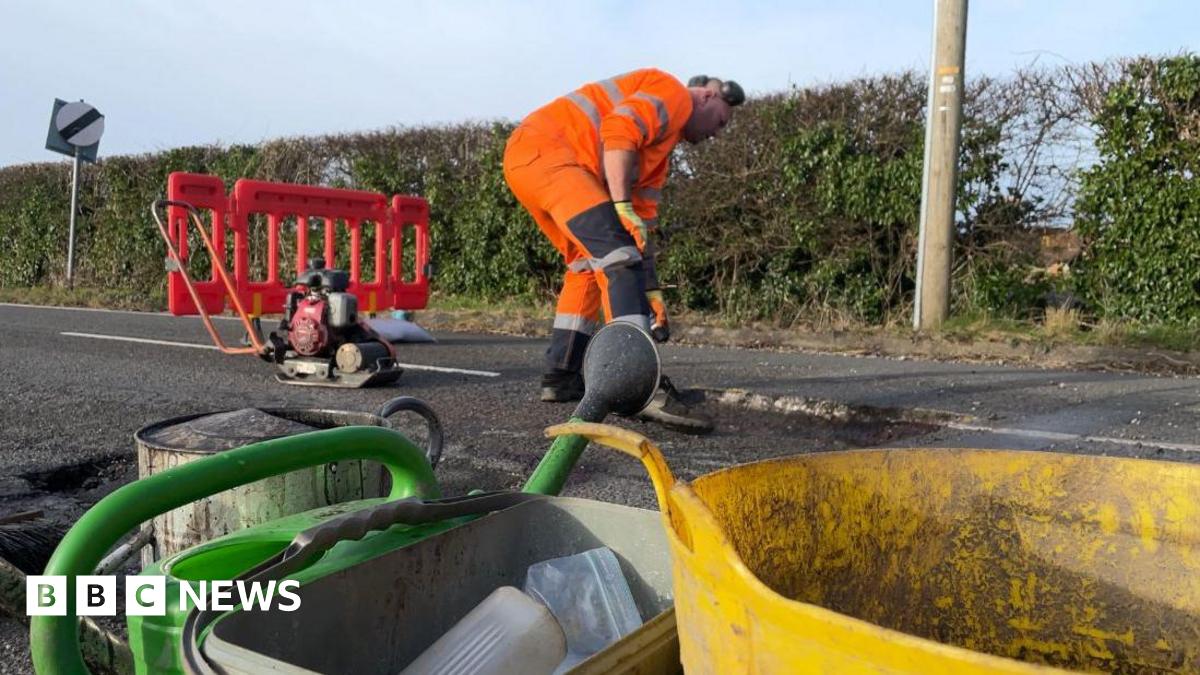 Road maintenance worker in orange jacket filling in a pothole on a road next to a hedge