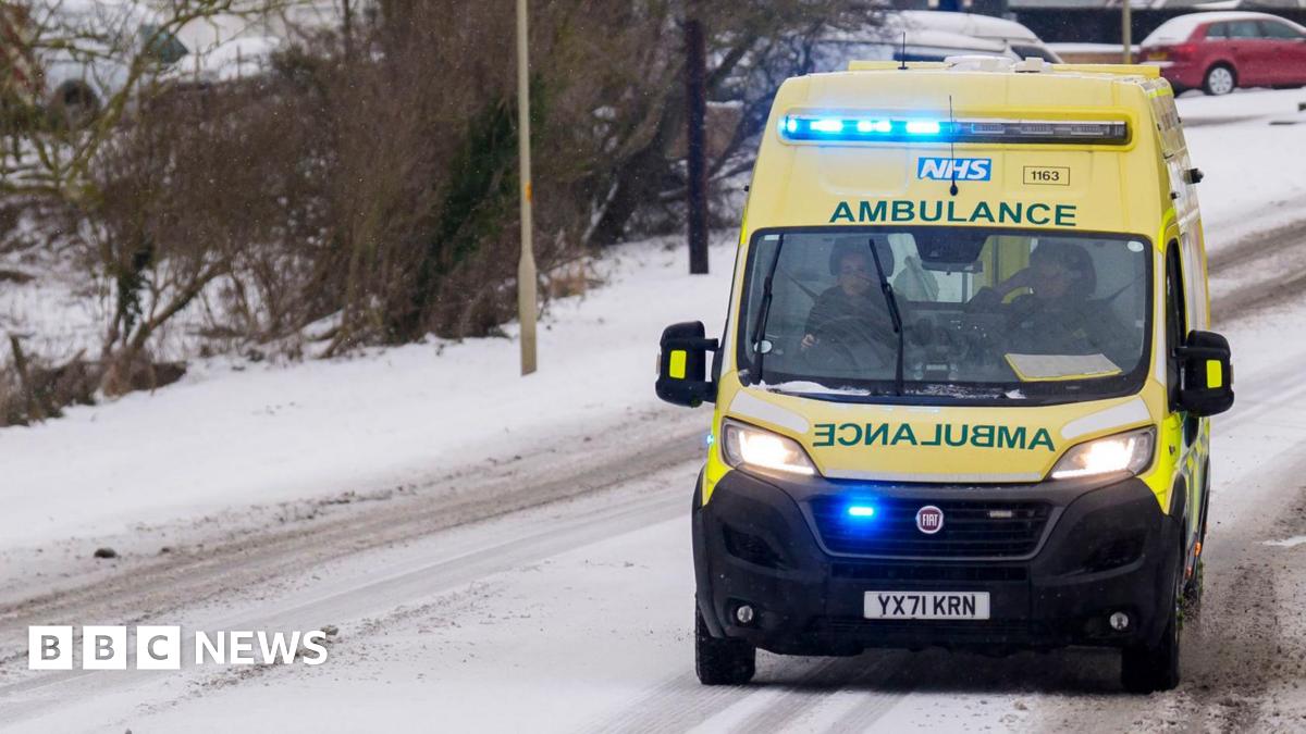 A yellow ambulance with its blue emergency lights on is driving along a snowy road. The grey tarmac is only peeking through where other cars have driven along it.