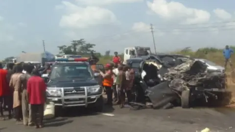 Federal Road Safety Corps A picture of a crashed black SUV beside an emergency vehicle and a crowd of people