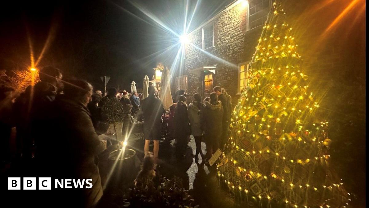 A group of people stand around a decorated Christmas tree outside, illuminated by lights and a single street lamp.