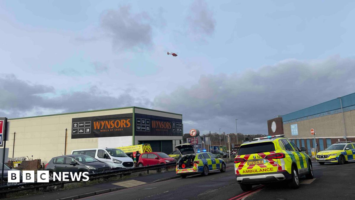 Emergency services parked either side of a road. There is a car park behind and a large Wynsors store with orange and black signange. There is an air ambulance helicopter above the scene.