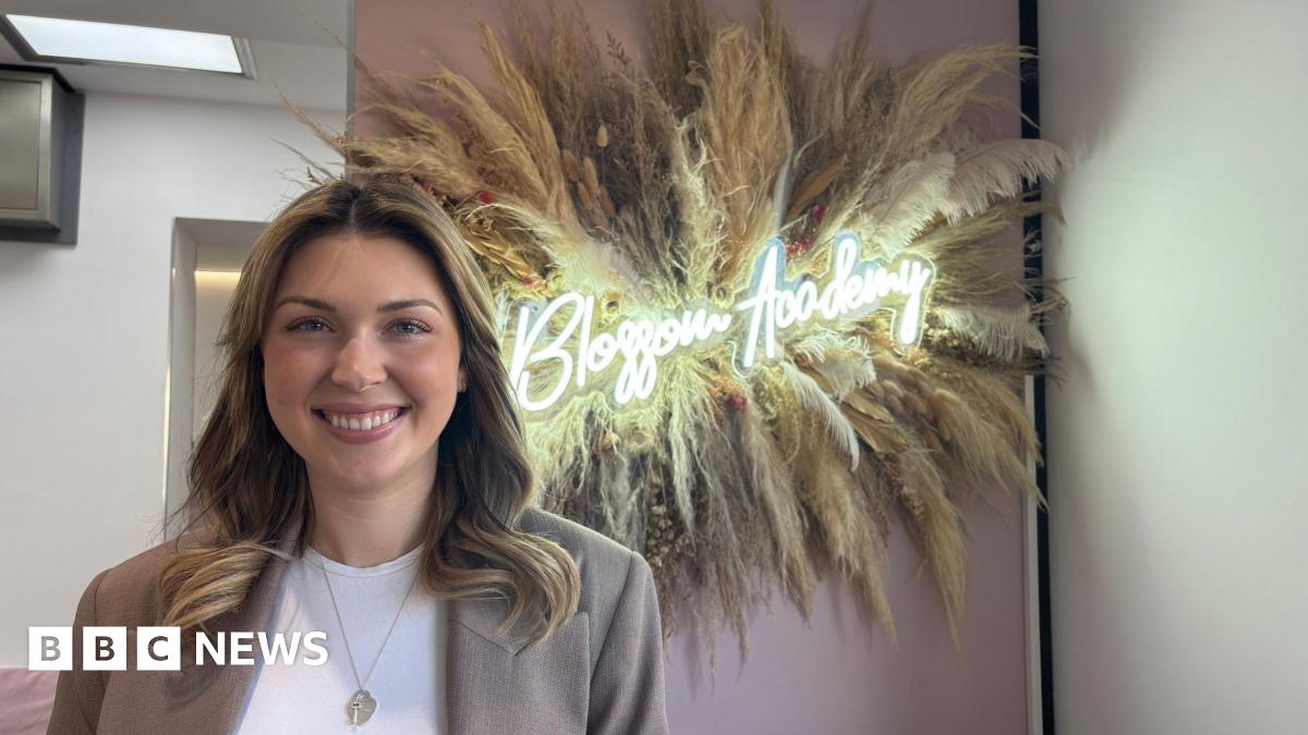 A woman with blonde and brown hair, wearing a white top and light brown blazer. She is standing in front of a pink wall, with a large display of pampas branches. There is a light in the middle that reads "Blossom Academy"