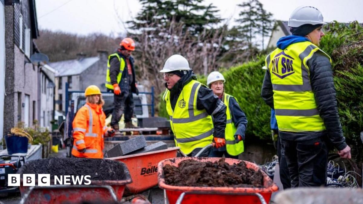 A group of construction workers in hard hats and high vis, They are surrounded by red wheelbarrows filled with dirt. On one side is  a bush and behind them are rows of grey houses.