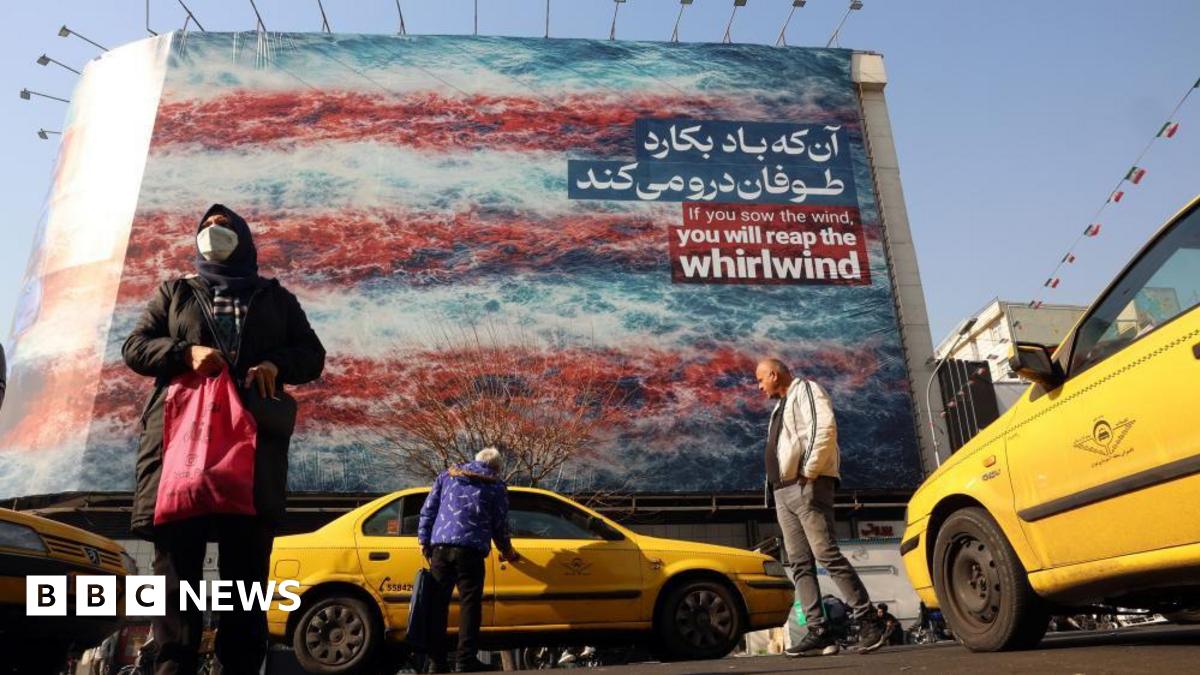 A woman is seen carrying a shopping bag and a man is seen getting into a yellow taxi cab in the foreground of the photo, while in the background there is a huge anti-US billboard which reads 'If you sow the wind, you will reap the whirlwind'