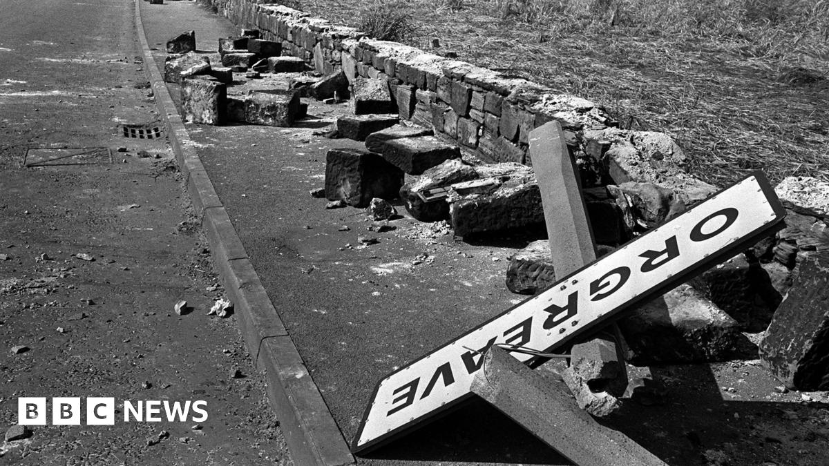 A black and white photo from 1984 of the Orgreave town road sign on the floor in a pile of rubble. A wall next to a road is torn down and the sign in lying on stones from the broken wall.