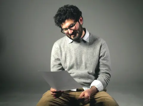 A man with thick, curly, dark hair, moustache and beard looks at a piece of paper. He is wearing glasses, a light grey jumper over a light shirt, and light brown trousers. 