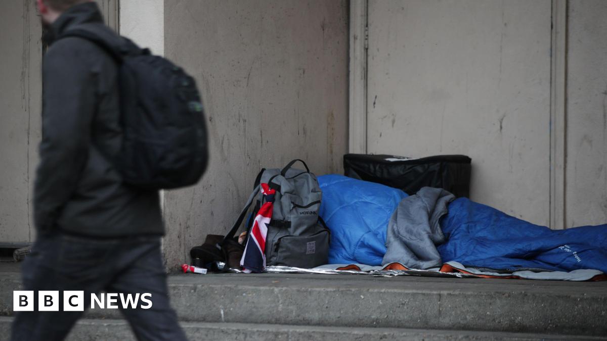 A person is sleeping in a doorway and is covered by a blue sleeping bag. A backpack is on the floor beside them. It appears to have a Union Jack flag tied around one of the loops on the bag. A pedestrian is walking past wearing a backpack.