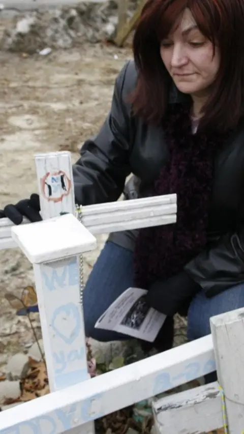 Gina visits the grave of her former fiance, Fred. She kneels down and has one hand on the white cross.