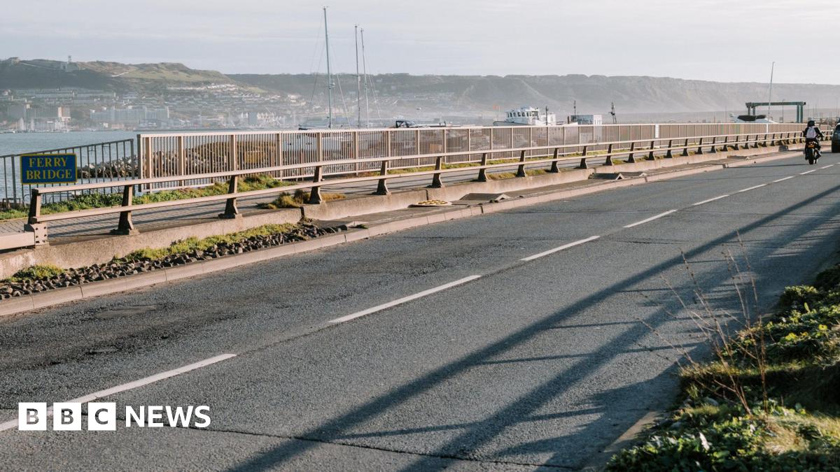 A grey road with white lines and grey fencing with green bushes either side and a number of boats in the background.