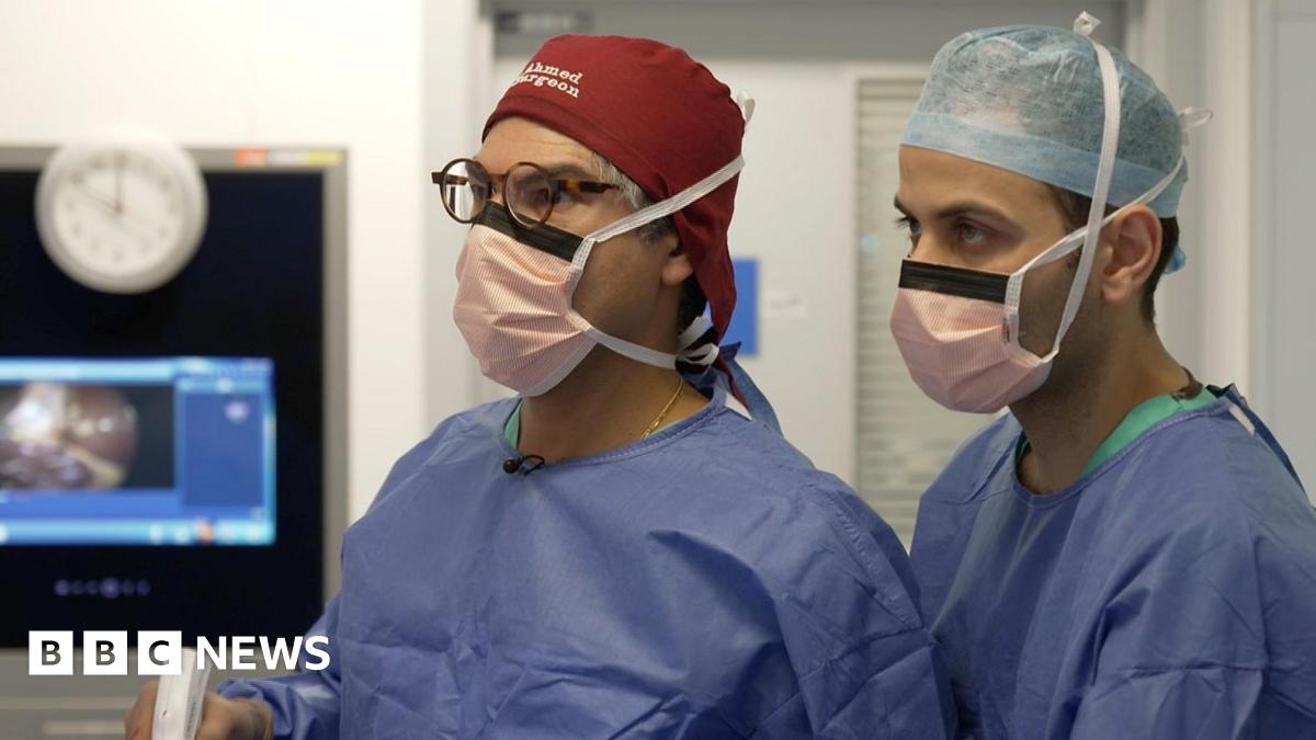 Two male surgeons in blue scrubs and face masks operate on a patient in a hospital. One of the surgeons is holding a white instrument in his hand. Behind them a screen in showing what they are operating on, but this is blurred.