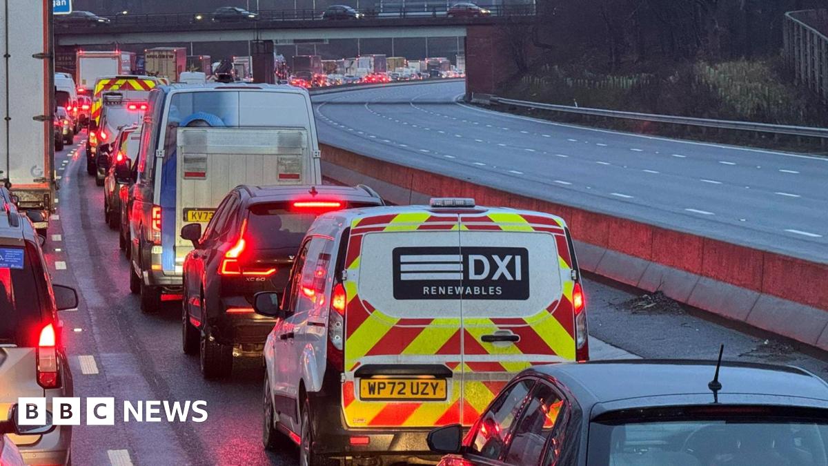 A long line of traffic visible along the M6 motorway, showing cars, vans and lorries with their rear brake lights illuminated red. The road curves to the right in the distance.