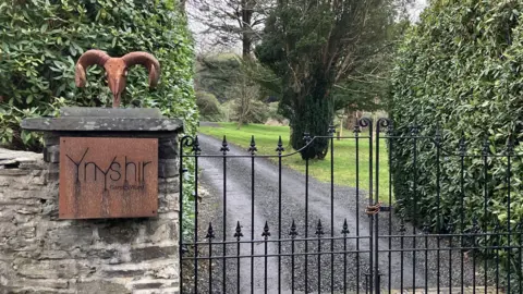 BBC The gated entrance to Ynshir restaurant. A black cast iron gate leads up a drive. On the wall is a bronze coloured sign with the restaurant's name on it.  
