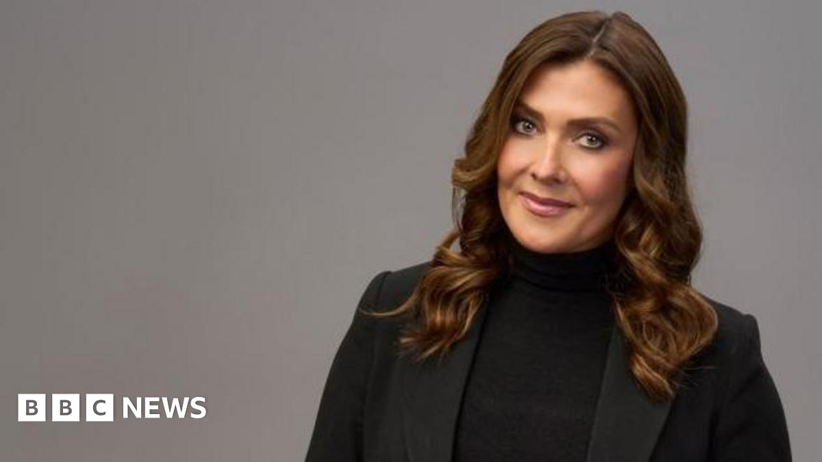 Kym Marsh, who is wearing a black blazer and black polo neck jumper, is standing in front of plain grey background for this posed photograph and is smiling.