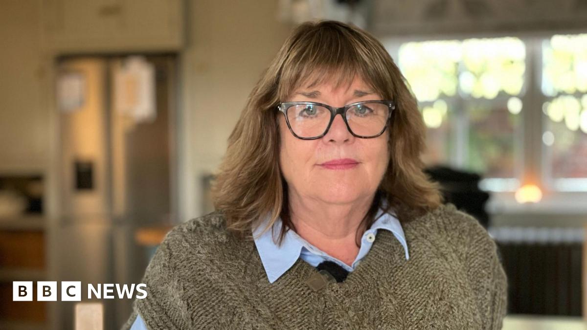 A woman with glasses and dark hair, wearing a brown knitted jumper is sitting indoors in front of a window.