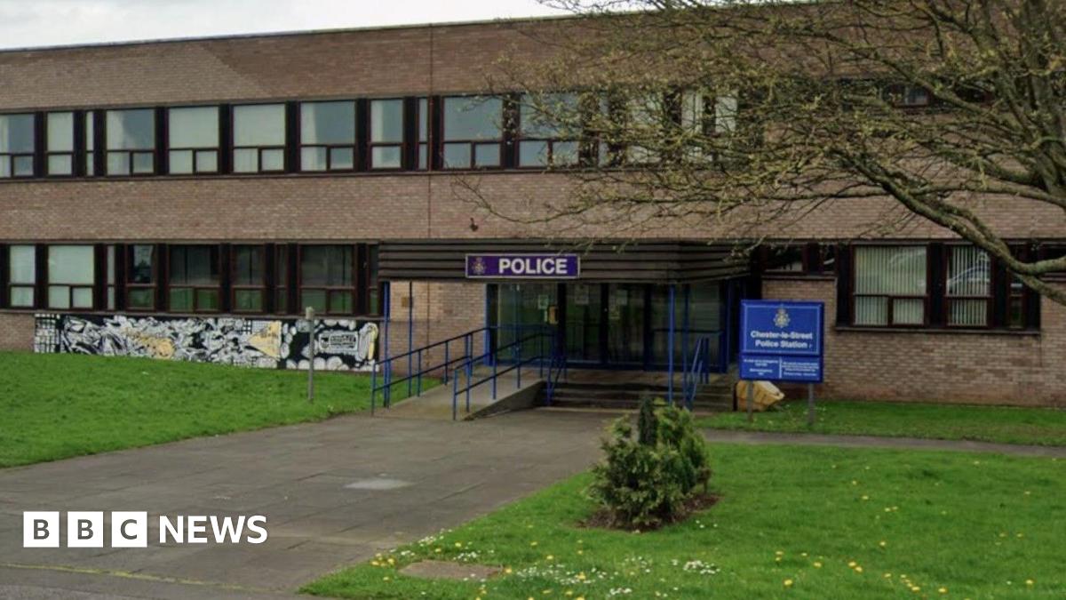 The outside of Chester-le-Street police station which is a large brown brick, brutalist-looking building which many windows with brown window panes. There is a blue sign which reads 'POLICE' above the entrance with a blue Chester-le-Street Police Station sign to the right. There is grass on either side of the entrance.