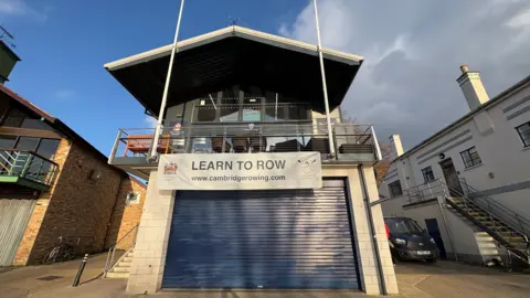 Ben Schofield/BBC A building, centre frame and shot from below. It is a boathouse on the River Cam, though the water is not in frame. It has a blue, roller shutter type door, over which a banner reading "learn to row' is hung. An external staircase can be just be seen on the left. On the first floor is a balcony with chairs. Two white flag poles stretch up from the balcony railings. The building has a pitched roof, which is set against a mostly blue sky, though there are some clouds. There are similarly sized buildings on either side.