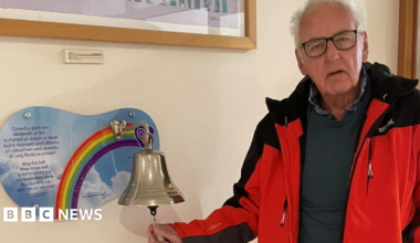 Noel Thomas looks at the camera. He has short white hair and has black-rimmed glasses. He is wearing a blue jumper with a black gilet. A dining table chair can be seen behind him as well as a door and a blue wall. It is a head and shoulders shot.