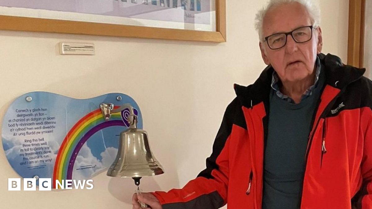 Noel Thomas looks at the camera. He has short white hair and has black-rimmed glasses. He is wearing a blue jumper with a black gilet. A dining table chair can be seen behind him as well as a door and a blue wall. It is a head and shoulders shot.