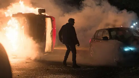 ITV The figure of a character standing in silhouette on a road at night in front of a burning coach and a crashed car, in a scene from Corriedale