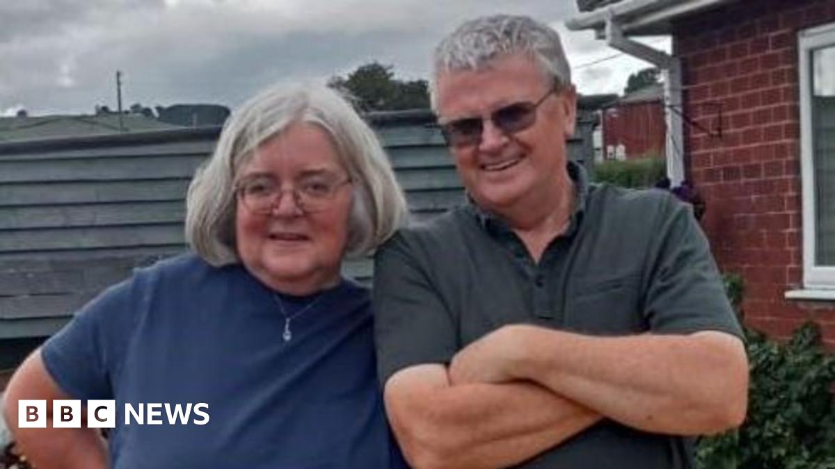A woman with bobbed grey hair and a man with short grey hair are smiling while standing next to each other. They are on decking, with railing and a house behind them.