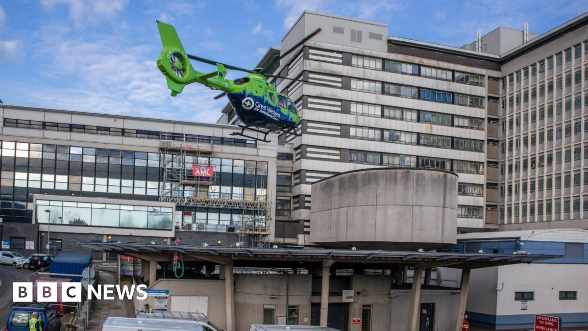 Great Western Air Ambulance's green and blue helicopter landing at a hospital. The building is several storeys high and has lots of vehicles parked outside.