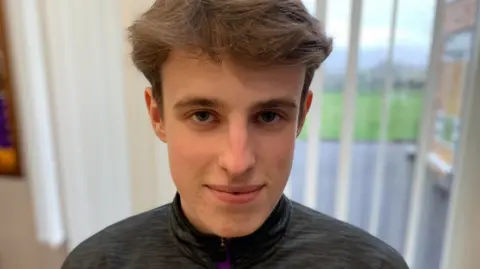 Cormac has brown hair and is smiling at the camera. He is standing in front of a window. He is wearing a grey, yellow and purple zip up top.