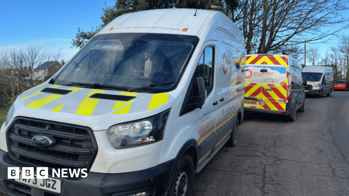 Two Wales and West vans parked on a roadside. They are mostly white but one has diagonal yellow stripes on its bonnet and the other has red and yellow chevrons on its back doors