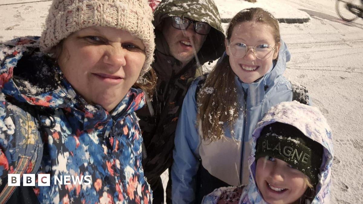 A selfie picture of a woman in a beige woolly hat and a red black and white patterned jacket. She is scrunching up her nose and mouth to the camera, as if in disgust. Crouched behind her is a man in glasses wearing a green jacket with the hood up, a smiling girl with glasses, long blonde hair and a blue jacket, and a shorter girl with a black hat and a light-coloured hood up. They are standing in a snowy street.