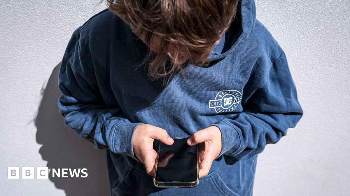 A boy in a blue hoodie looks down at his black phone he is holding in his hands, the screen is blank