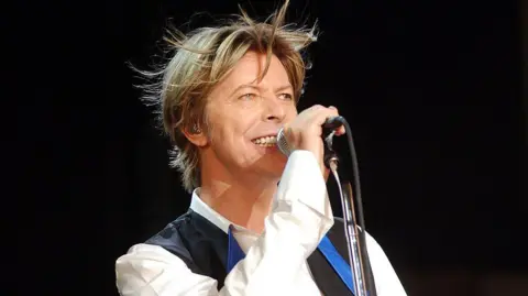 Getty Images A colour photograph of a David Bowie performing on stage in 2003, holding a microphone and smiling, wearing a white shirt and dark waistcoat under stage lighting.